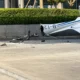 Small white airplane wrecked on a road with blue stripes tail damaged and scattered debris nearby
