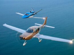 Two small private airplanes flying in formation over a calm blue ocean with a tan aircraft in the foreground and a blue aircraft behind it