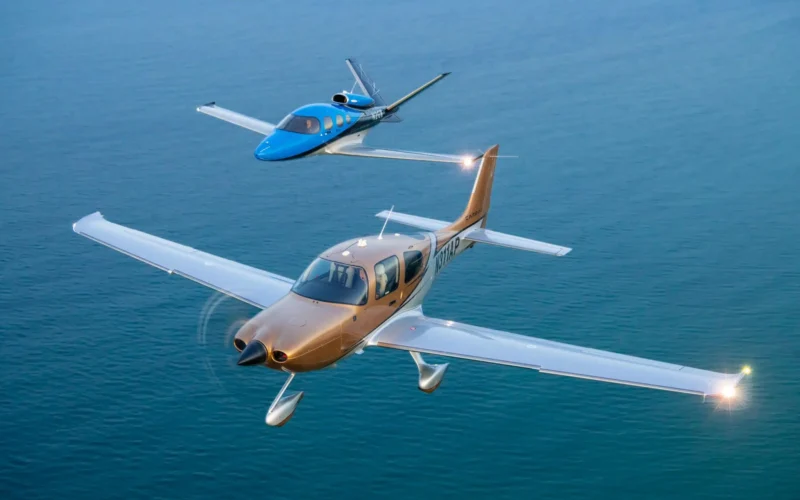 Two small private airplanes flying in formation over a calm blue ocean with a tan aircraft in the foreground and a blue aircraft behind it