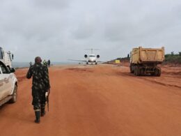 A soldier walks down a red dirt airstrip toward a small white jet with trucks parked on both sides