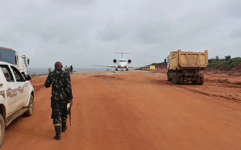 A soldier walks down a red dirt airstrip toward a small white jet with trucks parked on both sides