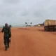 A soldier walks down a red dirt airstrip toward a small white jet with trucks parked on both sides