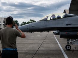 Ground crewman with headset facing a gray fighter jet on the tarmac cockpit with two pilots visible inside the canopy