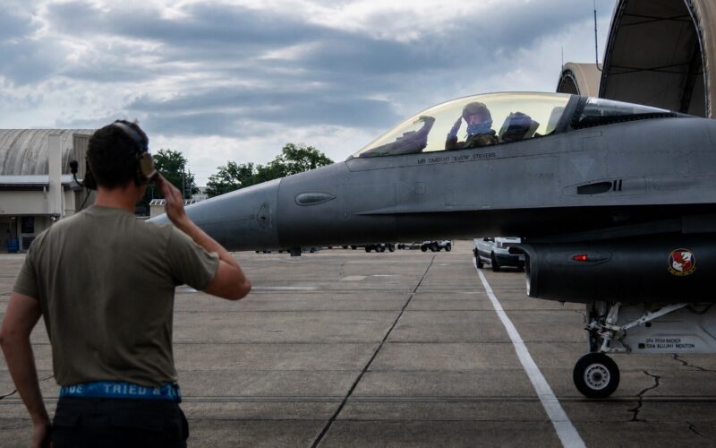 darpaf16 Ground crewman with headset facing a gray fighter jet on the tarmac cockpit with two pilots visible inside the canopy