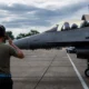 Ground crewman with headset facing a gray fighter jet on the tarmac cockpit with two pilots visible inside the canopy