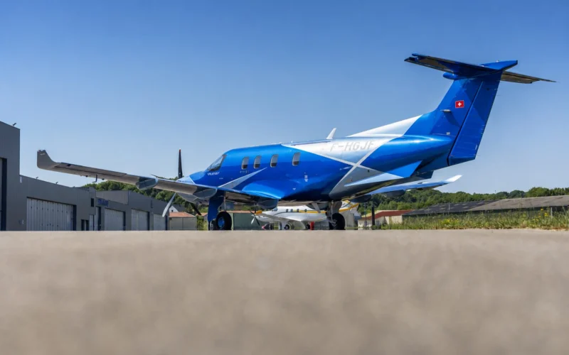 Blue private jet parked on a tarmac in front of airport hangars under a clear blue sky Swiss flag on tail left visible
