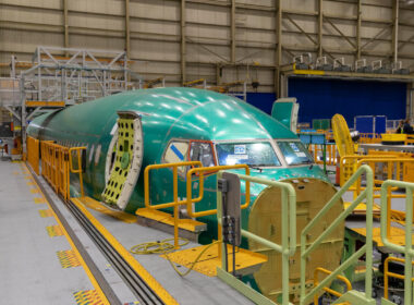 Green aircraft fuselage section on an assembly line inside a hangar, surrounded by yellow safety rails and equipment.