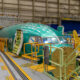 Green aircraft fuselage section on an assembly line inside a hangar surrounded by yellow safety rails and equipment