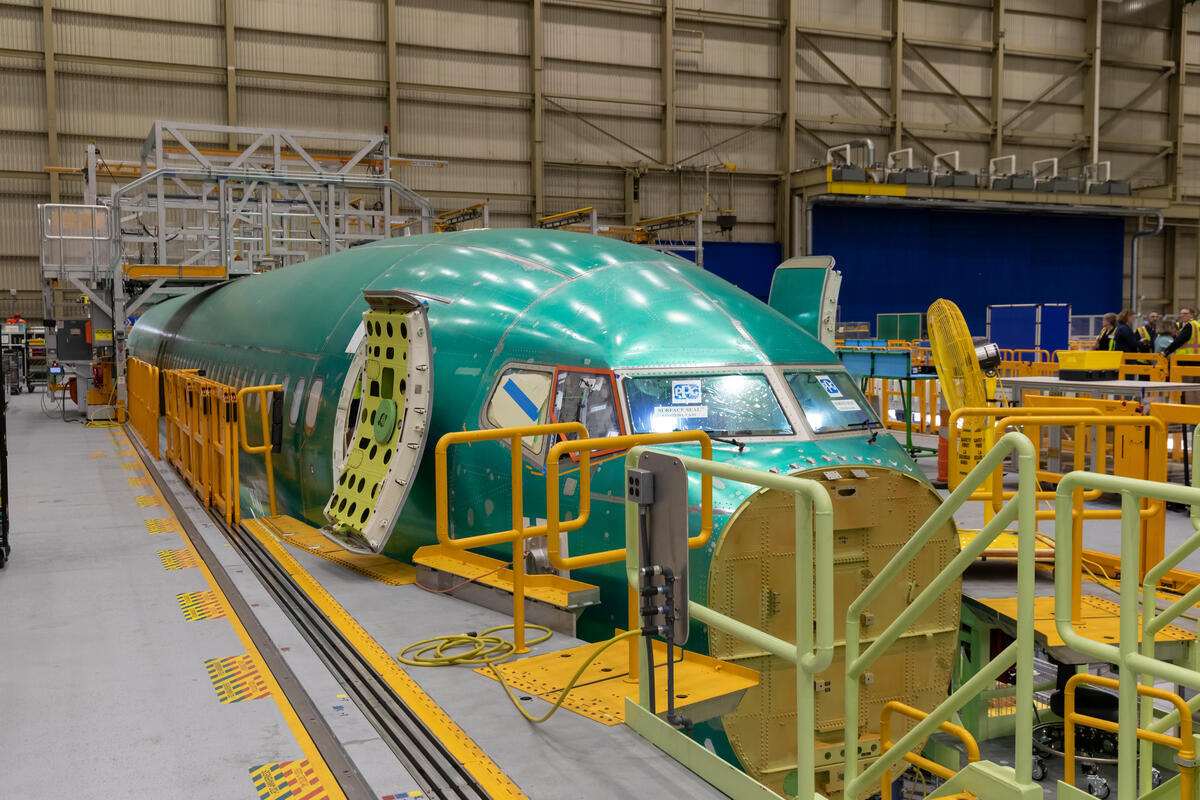 Green aircraft fuselage section on an assembly line inside a hangar surrounded by yellow safety rails and equipment