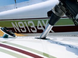 Close up of a small airplane wing and landing gear area showing a silver support strut passing through an opening in the wing with a coiled chain and hardware in the background
