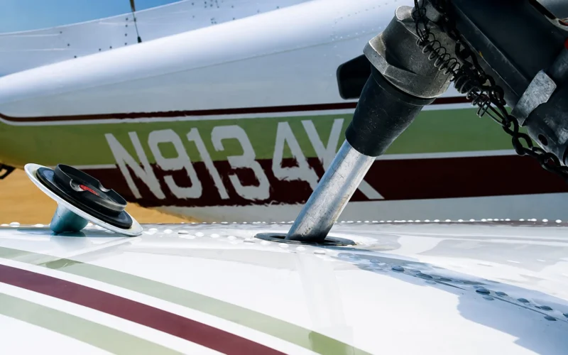Close up of a small airplane wing and landing gear area showing a silver support strut passing through an opening in the wing with a coiled chain and hardware in the background