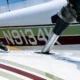 Close up of a small airplane wing and landing gear area showing a silver support strut passing through an opening in the wing with a coiled chain and hardware in the background