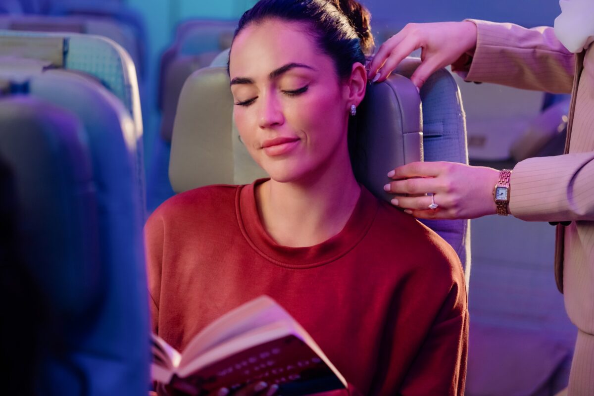 image Woman with eyes closed reading a book while a person pets her head on an airplane seat armrest nearby