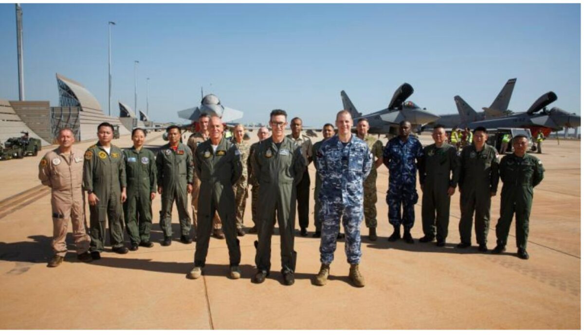 image Group of diverse military personnel in flight suits standing on an airstrip with fighter jets in the background under a clear blue sky