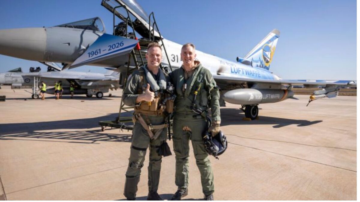 image Two pilots in flight suits pose with a fighter jet on a desert airstrip giving a thumbs up