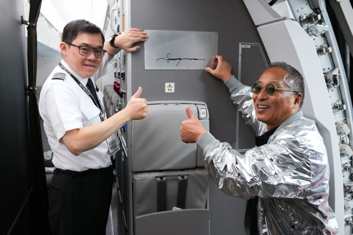 image Two men in an airplane galley pose and give thumbs up one in uniform and the other in a shiny silver jacket beside a large cabinet door
