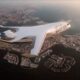 Futuristic white Gulf Air aircraft flying over a coastal city with a harbor and high rise skyline at sunset