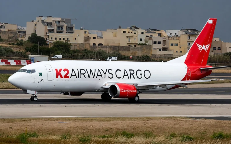 Cargo airplane with K2 AIRWAYS CARGO branding taxiing on a runway near beige buildings in the background overcast sky