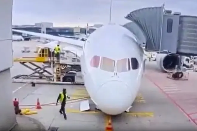 Front view of a white passenger airliner at the gate jet bridge connected and ground crew nearby on the tarmac