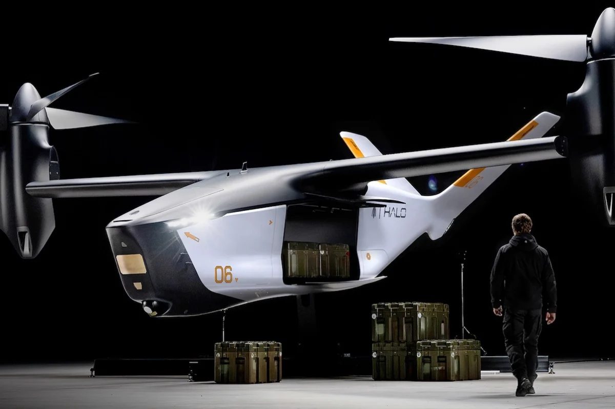 modular payload White futuristic twin rotor aircraft in a dim hangar with cargo crates beneath and a person in dark gear nearby