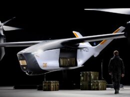 White futuristic twin rotor aircraft in a dim hangar with cargo crates beneath and a person in dark gear nearby