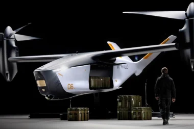 White futuristic twin rotor aircraft in a dim hangar with cargo crates beneath and a person in dark gear nearby