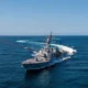 Gray US Navy destroyer sails through deep blue ocean under a clear sky American flag fluttering on the hull