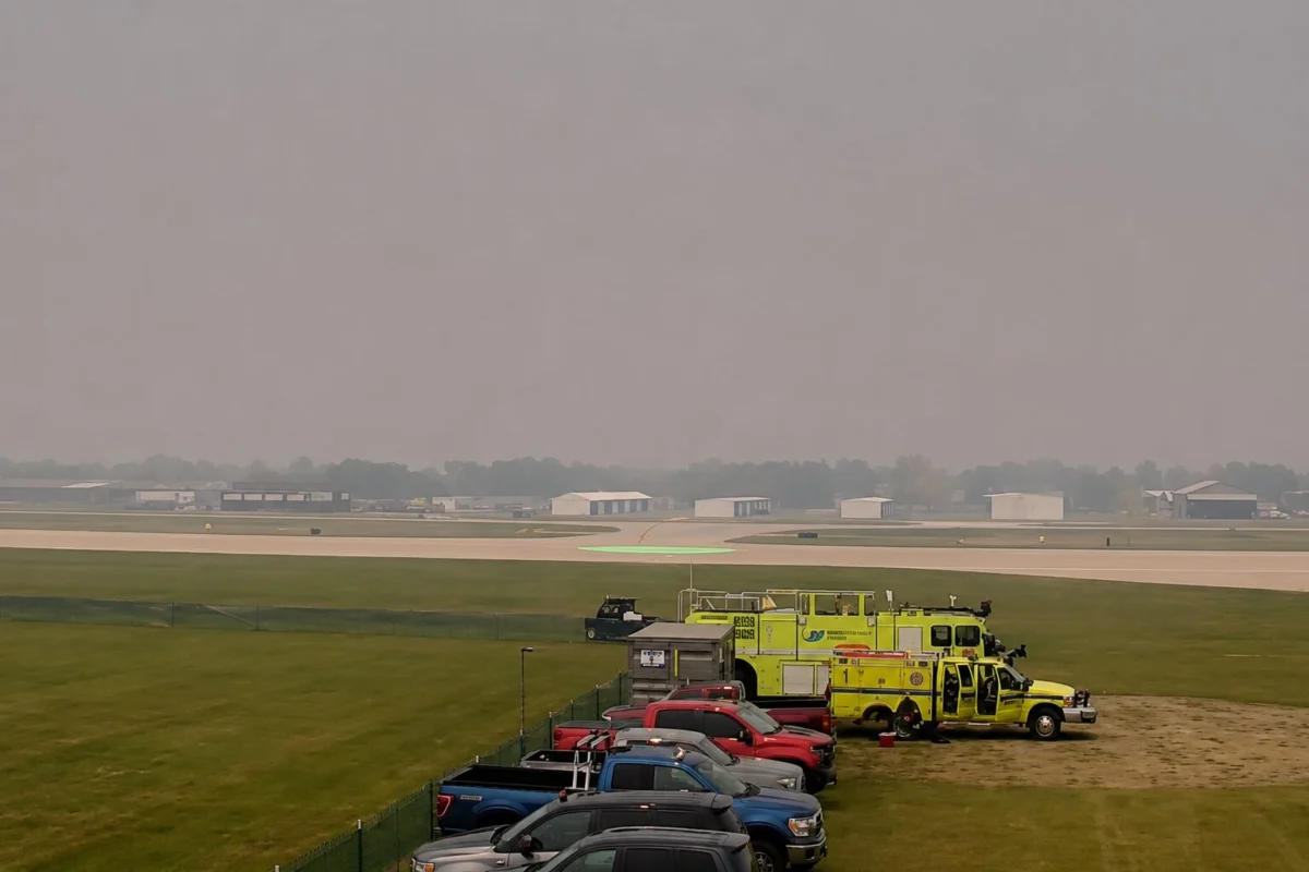 oshsmokey Line of parked cars and bright yellow emergency vehicles beside a runway at a hazy airport A green circular helicopter pad is visible in the distance