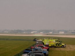 Line of parked cars and bright yellow emergency vehicles beside a runway at a hazy airport A green circular helicopter pad is visible in the distance
