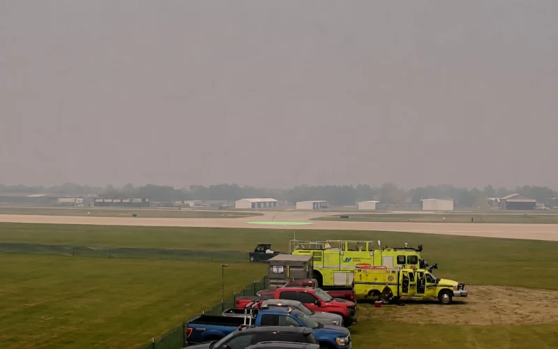 Line of parked cars and bright yellow emergency vehicles beside a runway at a hazy airport A green circular helicopter pad is visible in the distance