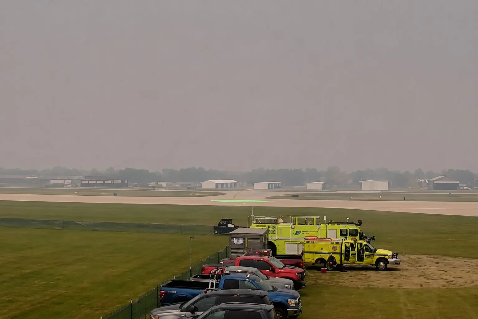 Line of parked cars and bright yellow emergency vehicles beside a runway at a hazy airport A green circular helicopter pad is visible in the distance