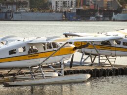Seaplanes with a yellow stripe tied to a dock on calm water Kenmore Air branding visible along the fuselage with a harbor backdrop in the distance