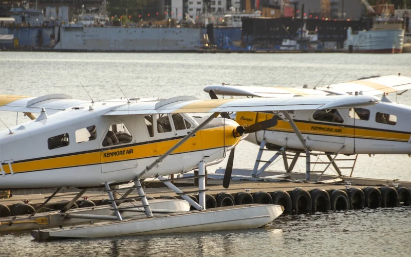 Seaplanes with a yellow stripe tied to a dock on calm water Kenmore Air branding visible along the fuselage with a harbor backdrop in the distance
