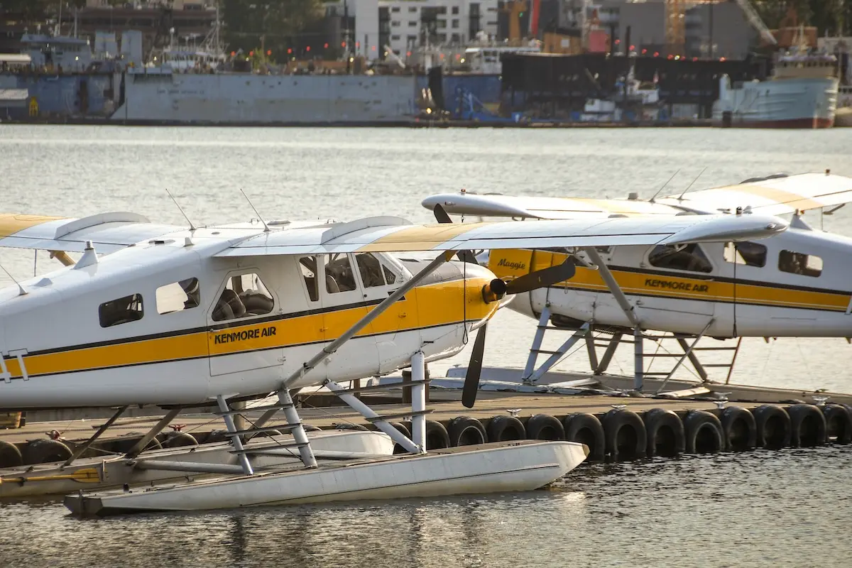 SeattleWaUsa June2018FloatplanesOperatedByKenmore Seaplanes with a yellow stripe tied to a dock on calm water Kenmore Air branding visible along the fuselage with a harbor backdrop in the distance