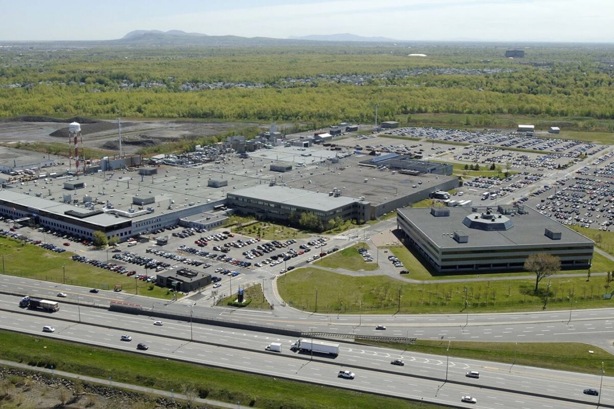 Aerial view of a large office campus with multiple flat roofed buildings extensive parking lots and surrounding roads near a wooded area and distant hills