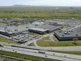 Aerial view of a large office campus with multiple flat roofed buildings extensive parking lots and surrounding roads near a wooded area and distant hills