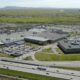 Aerial view of a large office campus with multiple flat roofed buildings extensive parking lots and surrounding roads near a wooded area and distant hills