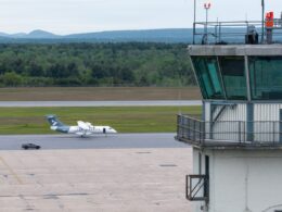 Airfield scene with a control tower on the right a small white aircraft on the taxiway and a service vehicle nearby with grassy fields and distant hills in the background