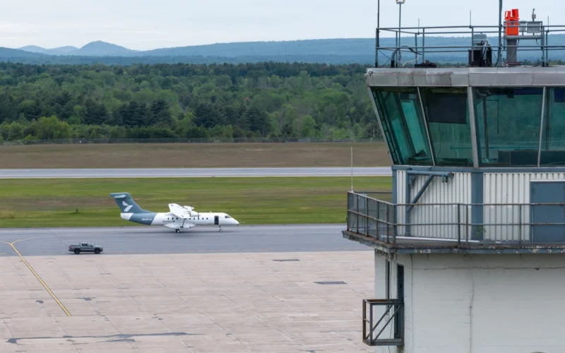 Airfield scene with a control tower on the right a small white aircraft on the taxiway and a service vehicle nearby with grassy fields and distant hills in the background