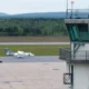 Airfield scene with a control tower on the right a small white aircraft on the taxiway and a service vehicle nearby with grassy fields and distant hills in the background