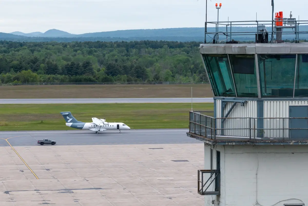 Airfield scene with a control tower on the right a small white aircraft on the taxiway and a service vehicle nearby with grassy fields and distant hills in the background