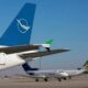 Airplanes parked on a sunny tarmac with a large blue tail fin in the foreground and a Syrian Airlines jet in the midground behind it