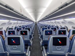 Interior of a modern passenger jet cabin with rows of blue seats and individual in flight entertainment screens along the backs of the seats viewed down the aisle from the rear