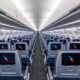 Interior of a modern passenger jet cabin with rows of blue seats and individual in flight entertainment screens along the backs of the seats viewed down the aisle from the rear