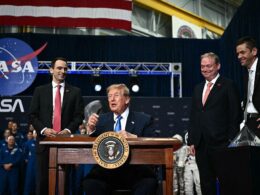 Donald Trump speaking at a podium with the Presidential Seal and NASA backdrop flanked by two suited men as astronauts and staff look on