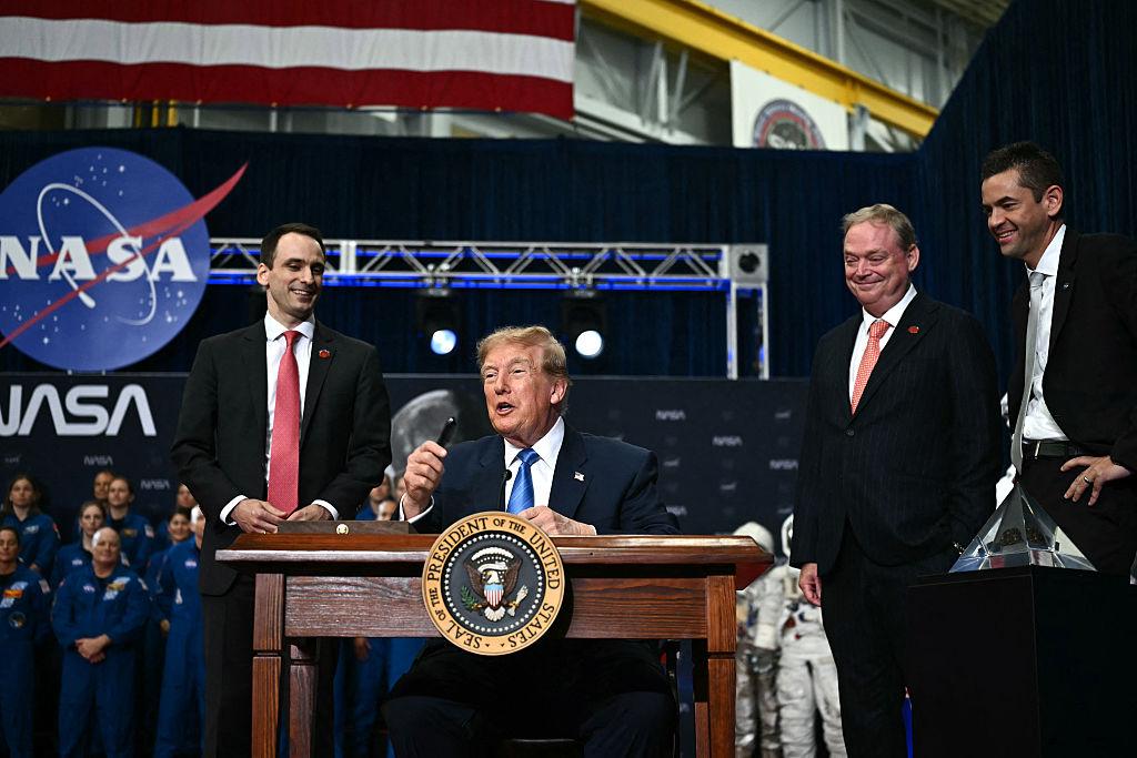 Donald Trump speaking at a podium with the Presidential Seal and NASA backdrop flanked by two suited men as astronauts and staff look on