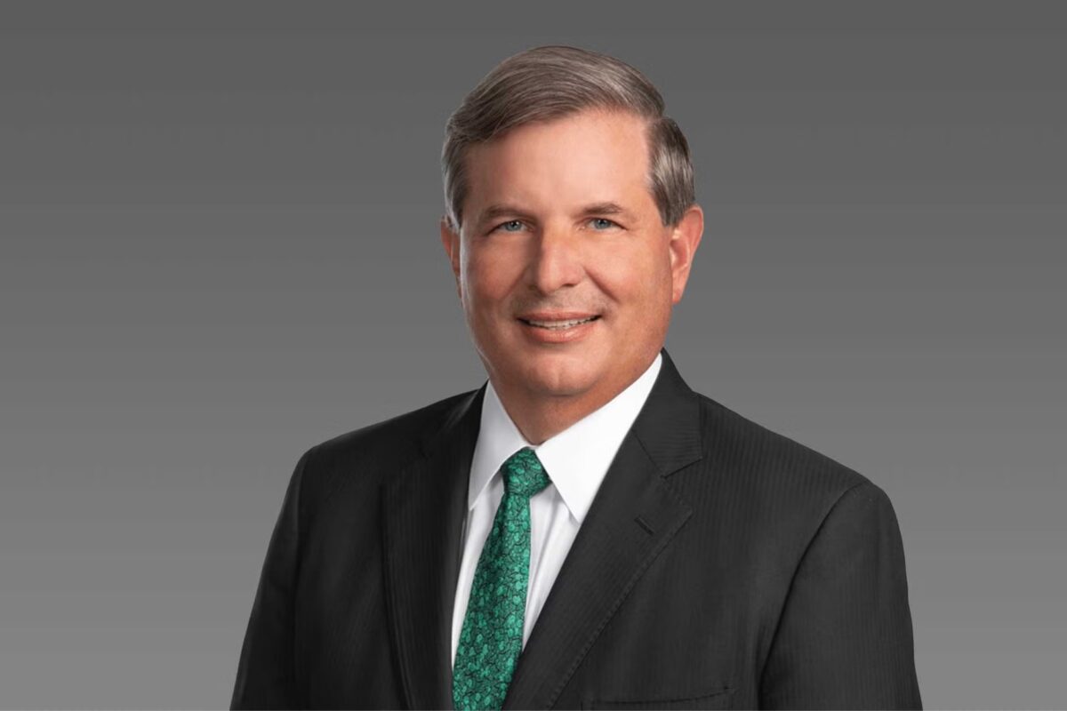Professional headshot middle aged man in a dark suit and white shirt with a green patterned tie smiling against a gray gradient background