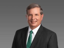 Professional headshot middle aged man in a dark suit and white shirt with a green patterned tie smiling against a gray gradient background