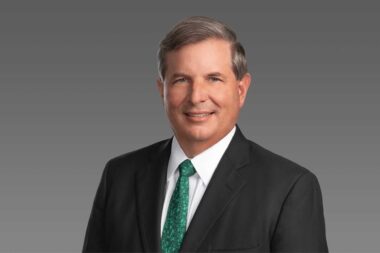 2022 CEO chris kubasik green Professional headshot middle aged man in a dark suit and white shirt with a green patterned tie smiling against a gray gradient background
