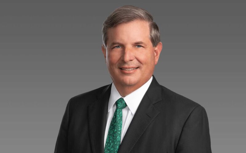 2022 CEO chris kubasik green Professional headshot middle aged man in a dark suit and white shirt with a green patterned tie smiling against a gray gradient background